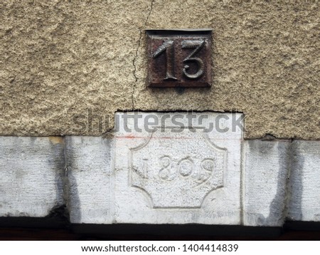 House number thirteen (13) wooden textured sign brown plate against off-white cracked stone wall background and above 1809 AD exact year inscription on rough brick (France). Grunge, texture, wallpaper