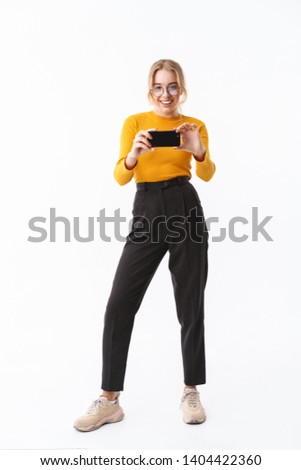 Full length of an attractive young girl wearing sweater standing isolated over white background, taking a picture with mobile phone