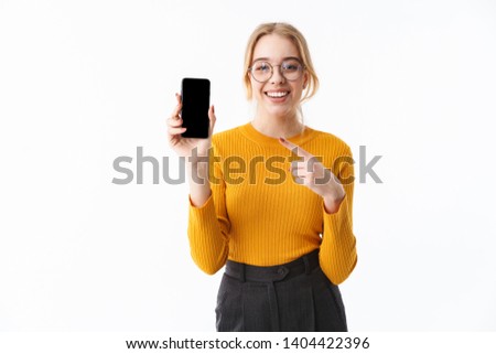 Attractive young girl wearing sweater standing isolated over white background, pointing finger at blank screen mobile phone