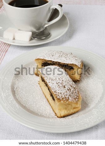 Bun filled with poppy seeds, cut into halves, sprinkled with sugar, served on the white plate with a cup of coffee, on the table covered with the table cloth.
