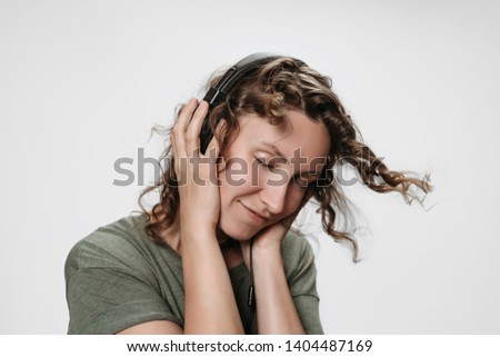 Carefree cheerful young curly woman listen favourite music with hand on her headphones, smiles positively, dance actively, moves to the music shakes curls, have fun, enjoy music. Isolated on white.