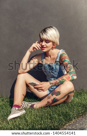 Young alternative girl sitting on grass isolated on grey wall on the city street using application on smartphone concentrated smiling joyful