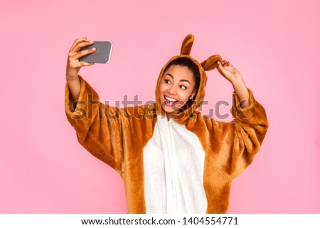 Young woman in bunny kigurumi standing isolated on pink background taking selfie photo on smartphone touching rabbit ears looking camera smiling playful
