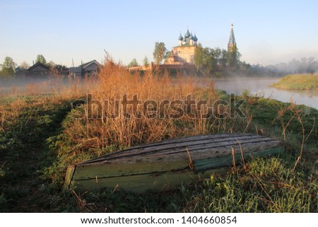 Old wooden turned boat on riverside in misty summer village morning with background of church and blue sky