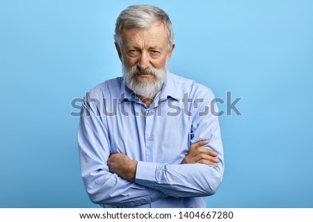 Handsome senior man in blue shirt with skeptic, nervous expression standing with crossed arms. close up portrait. isolated light blue background Royalty-Free Stock Photo #1404667280