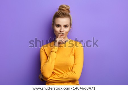 Attractive girl in casual clothes keeping hand on chin in doubt and suspicion, feeling sceptical about something, isolated blue background, studio shot