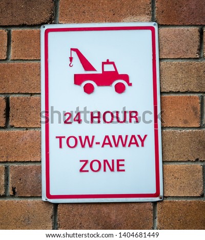 Old peeling red and white sign hanging on brown brick wall with text 24 HOUR TOW-AWAY ZONE and picture of tow truck