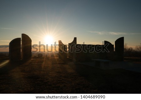 Gods new day a sunset with sunburst framed within the ten commandment tablets, an obelisk, a monolith with sun behind against a blue sky with clouds grass and rocks foreground all silhouetted 