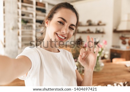 Image of charming optimistic woman wearing casual clothes taking selfie photo and showing ok sing in cozy kitchen