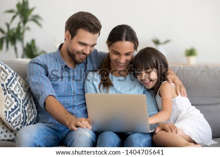 Smiling young parents have fun rest on sofa watching funny video on laptop with little girl child, happy family mom and dad relax on couch with preschooler daughter enjoy movie laughing at home