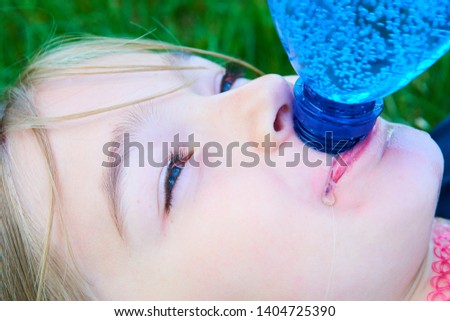 Close up portrait of adorable girl with blue eyes drinking water from plastic blue bottle with greenery background