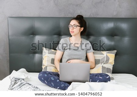 A beautiful young Asian woman in home clothes and glasses is sitting on the gray bed and holding a laptop, notebook. remote work