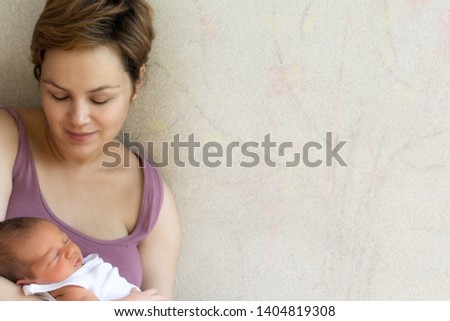 Young woman is holding her newborn son in her arms, watching him as he sleeps peacefully.