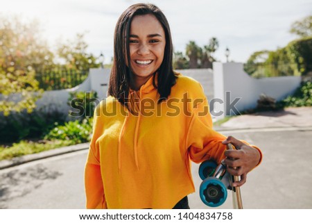 Cheerful teenage girl standing on street holding a skateboard. Smiling girl standing outdoors in the street on a sunny day.