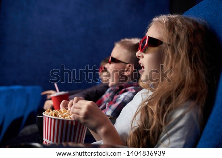 Shocked girl with opened eyes watching amazing film in movie house. Excited children wearing 3d glasses sitting together, eating tasty popcorn in cinema. Concept of leisure and fun.