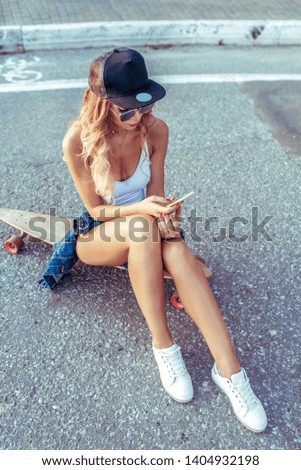 Girl skateboard summer city, her hand smartphone, writes message, reads online application to Internet. Denim sneakers baseball cap. Youth fashion style, modern leisure trend, tanned sports figure.