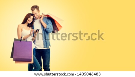 Photo of happy smiling young couple with shopping bags, and smartphone, over yellow color backround. Copy space for some slogan or advertising text. Caucasian models - online shopping concept.