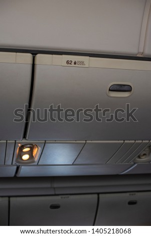 Closed up an airplane interior cabin, seeing an overhead storage compartment with number and seating position printed. Light for reading is on. 