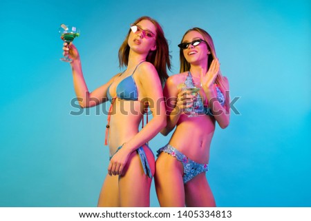 Beautiful young girls' half-length portrait isolated on blue studio background in neon light. Women posing in fashionable bodysuit. Facial expression, summer, weekend concept. Trendy colors.
