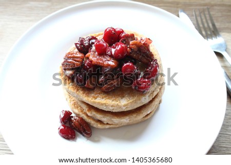 Gluten free buckwheat pancakes topped with cranberries and pecans glazed with maple syrup served on elegant round white plate and wooden surface, closeup 