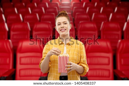 cinema, fast food and entertainment concept - smiling red haired teenage girl in checkered shirt eating popcorn from striped bucket over movie theater background