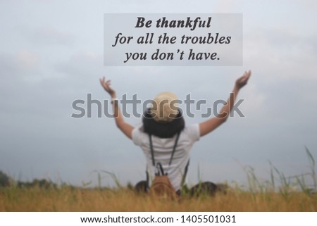 Inspirational quote- Be thankful for all the troubles you do not have. With blurry image of a young woman sitting from the back on the dried brown grass in the field, hand raised with open arms.
