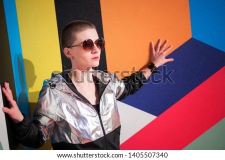 Portrait of young beautiful woman in sunglasses with short hair in a silver jacket on colourful geometric background, her hands touch the wall