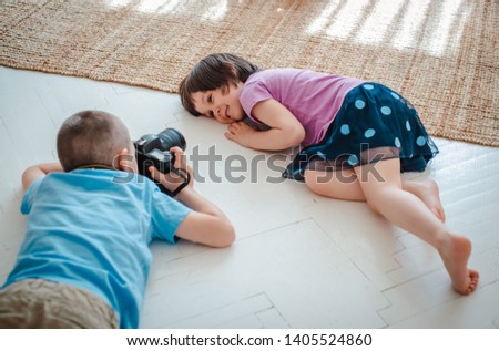 A small boy makes a portrait of a girl with a large camera in his hands. Brother photographs sister lying on floor in the room. Children's training profession reporter and photographer. Photo school