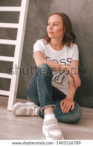 Photo of a young woman in a white t-shirt, jeans against a dark wall sitting on the floor