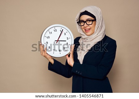  islam woman in a veil with glasses smiling keeps wall round clock on a beige background                             