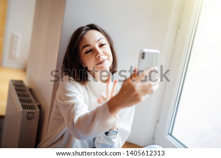 Pretty woman sitting on the windowsill and making selfie on smartphone