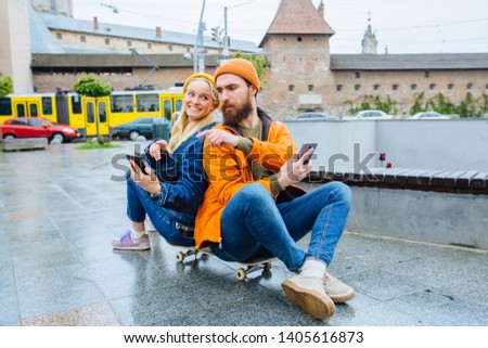 Teenage happy hipster stylish couple traveler looking at each other with smart phones while sitting on a longboard in rainy weather urban city street with traffic on background. Lifestyle concept.