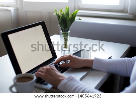 Mockup image of woman's hands using laptop with blank white screen on white table, coffee, tablet and a bouquet of lily of the valley. Woman's workplace in white colors. Home office. Copy space