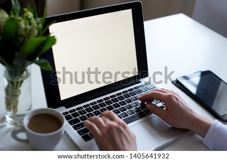 Mockup image of woman's hands using laptop with blank white screen on white table, coffee, tablet and a bouquet of lily of the valley. Woman's workplace in white colors. Home office. Copy space