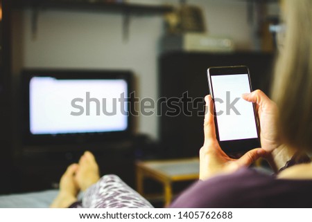 A woman with outstretched legs on the couch in the living room watches the TV and uses a DLNA on a smartphone, wireless internet, social media. Gambling, mobile betting. Addictions. Horizontal Royalty-Free Stock Photo #1405762688