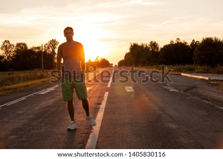 Young man or hipster in colourful vest and shorts stands on a free road with a beautiful sunset behind him