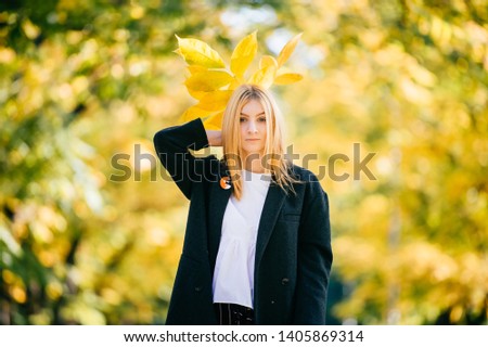 strange Young girl in black coat holding autumn leaves behind her head in nature on blurred yellow background. work of art by a romantic woman. lady in autumn in autumn Park. portrait on a belt