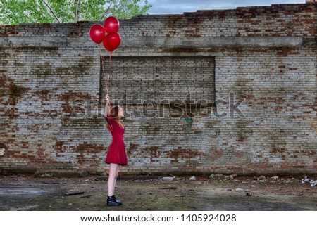 Beautiful young red hair girl in red dress with red helium balloons in hand inside of abandoned place with big ruined brick graffiti wall on background. HDR image