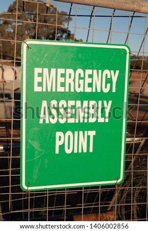 Emergency Assembly Point sign with white text on green, attached to wire frame fence, as featured on building or construction sites.
