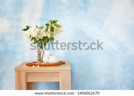Beautiful lilac flowers in vase with teapot on table against color background