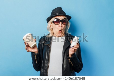 Old lady wearing glasses, a leather jacket and a hat is holding a glass with coffee and a bottle with pills on a blue background. Concept of abuse of relaxants, choice between health and harm
