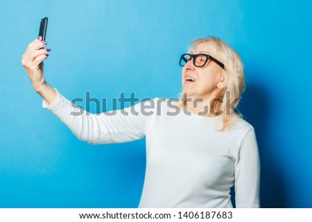 Old woman with glasses takes a selfie on a blue background. The concept of new technologies