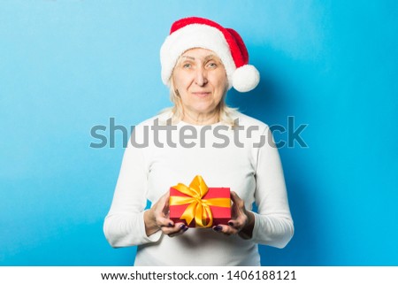 Old lady in a Santa Claus hat is holding a gift against a blue background. Concept of celebrating Christmas, new year, winter holidays, buying gifts