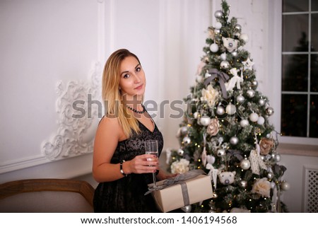 young woman in an elegant black dress sitting near the Christmas tree and drinking champagne

