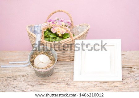 A white picture frame as a mock up - background - for creative ideas about Easter. Easter egg with nest in zinc bucket and basket of spring flowers Primroses stand on old wooden underground.