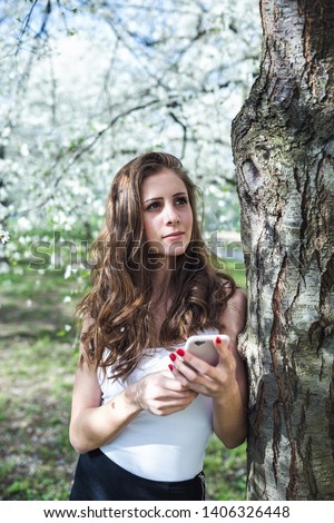 Young woman in a casual sport clothes holding a smart phone under the blooming cherry trees. Spring time.