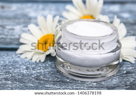 Cosmetic cream in a glass jar stands near chamomile flowers.