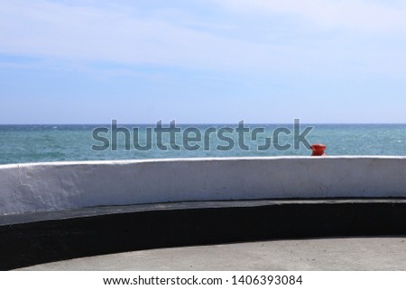 A bright red bollard is visible along a sea wall. The wall is white the sea is blue. Typical coastal image could be used in coastal holiday homes.