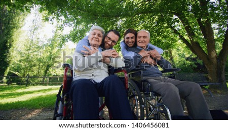 Portrait of happy grandchildren are hugging their grandparents both in a wheelchair as a sign of love and respect in a green park on a sunny day.
