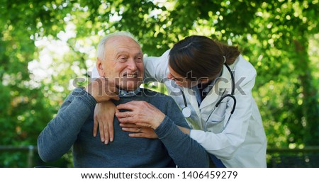 Portrait of carefree and happy young woman social worker or nurse is stroking the hand of senior man in a wheelchair as sign of care and support during they are having fun to talk to each other.